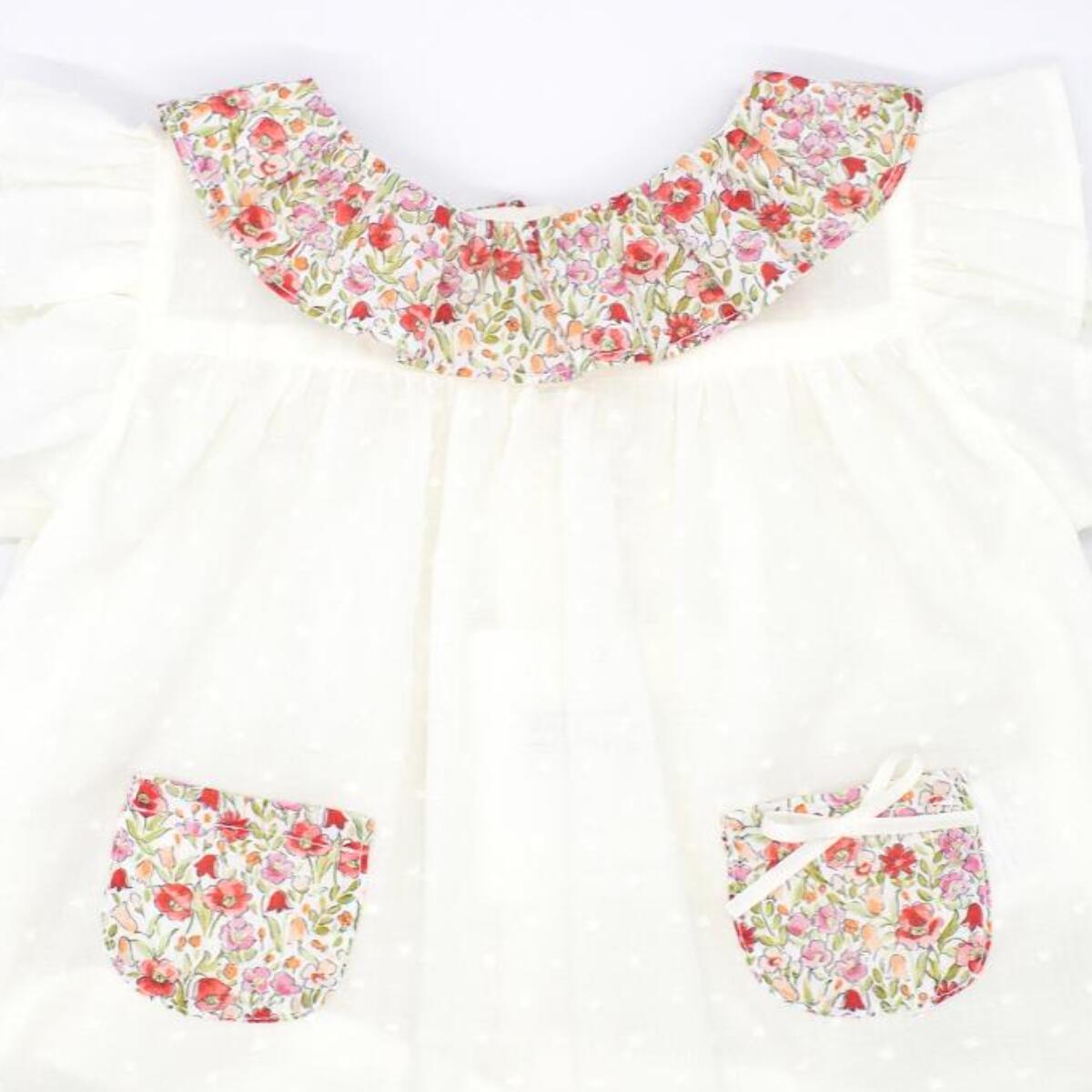 Blusa blanca con bolsillos y volante en el cuello de flores rojas, volantes en las sisas. Lazo en el bolsillo.