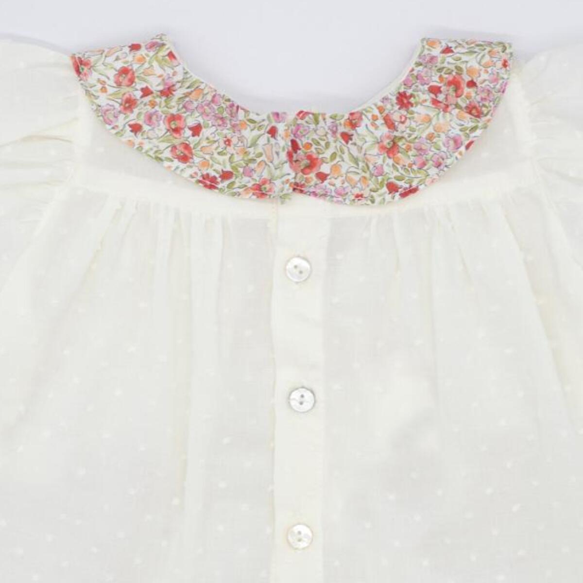 Blusa blanca con bolsillos y volante en el cuello de flores rojas, volantes en las sisas. Lazo en el bolsillo.