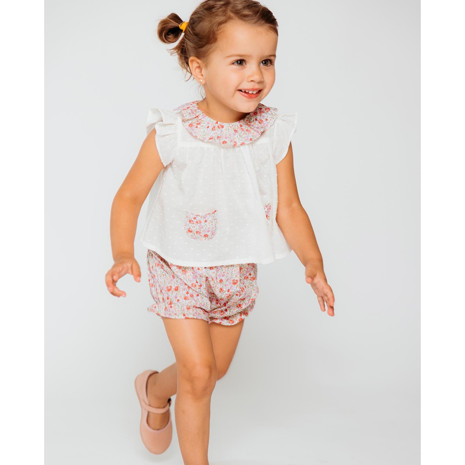 Blusa blanca con bolsillos y volante en el cuello de flores rojas, volantes en las sisas. Lazo en el bolsillo.