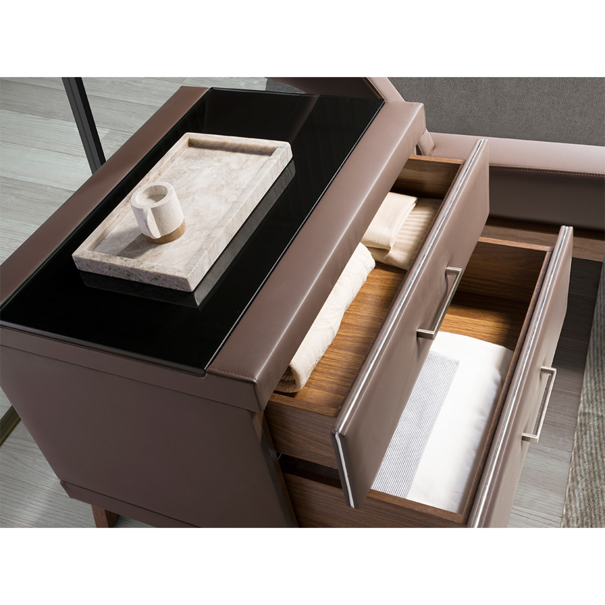 Mesita de noche Angel Cerdá con estructura de madera chapada en nogal natural tapizada en polipiel marrón chocolate y patas de madera chapada en nogal natural tapa de cristal templado tintado en negro 60x46x56cm