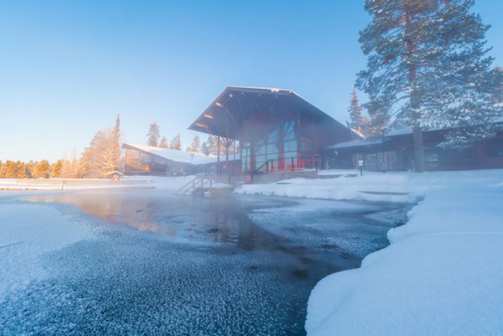 Edificio moderno sulla riva di un lago parzialmente ghiacciato e fumante in un paesaggio invernale innevato.